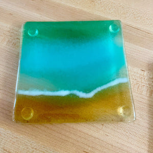 A fused glass coaster featuring a Turquoise green and gold beach scene is on a maple wood table background