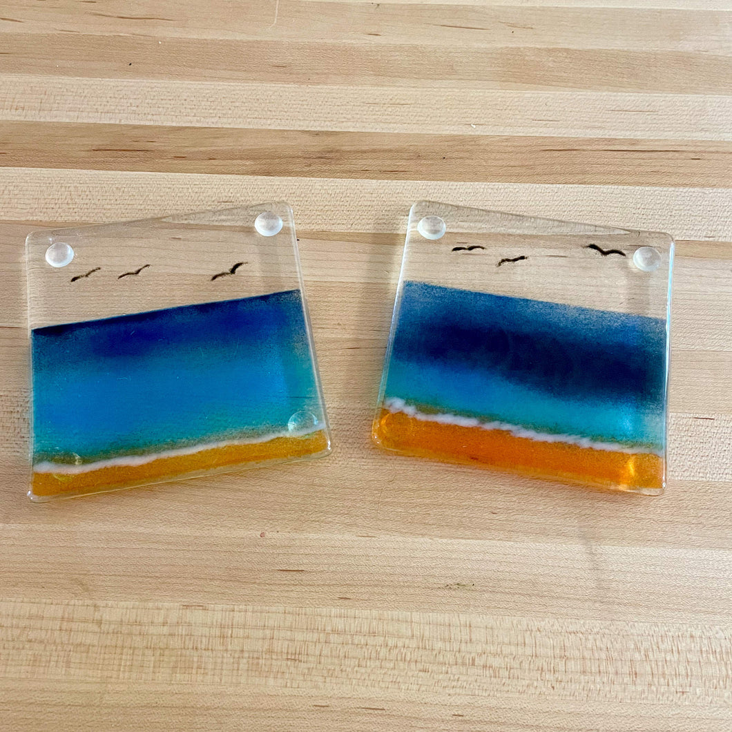 A pair of blue and gold beach scene coasters are on a maple table