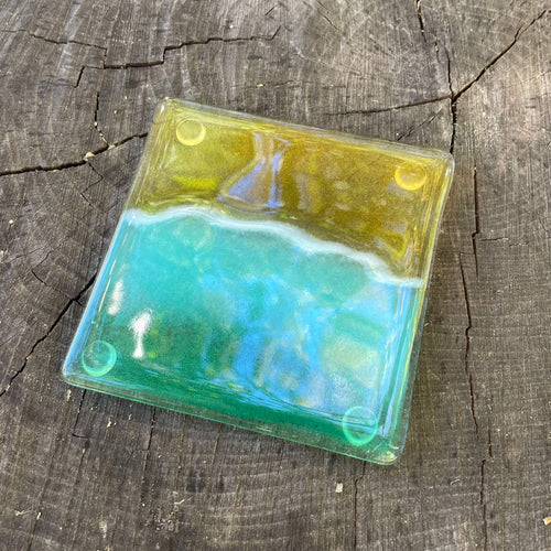 A fused glass coaster featuring a Turquoise green and gold beach scene is on a tree stump
