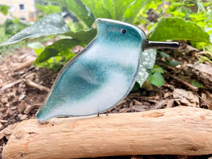 A fused glass bird-shaped kingfisher  bird with a black beak, standing on a light-colored log in an outdoor garden setting.