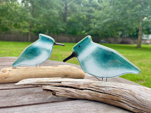 Two teal and white belted kingfisher bird figurines with black beaks, displayed on driftwood logs on a wooden table with a blurred green backyard background.