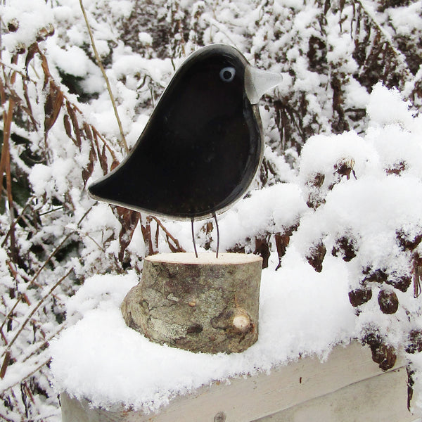 Load image into Gallery viewer, A black fused glass adult crow perched on a log in the garden/backyard in winter
