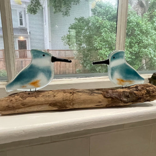  A pair of teal blue and white fused glass kingfishers perched on driftwood on a window ledge.