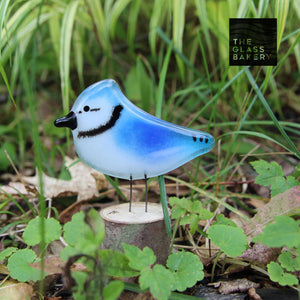 A small blue and white glass blue jay bird sits on a log in amongst green foliage.