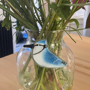 Clear glass vase with greenery and a blue fused glass hanging blue jay bird ornament on a wooden surface.