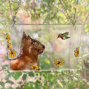 Decorative glass panel with a cat, hummingbird, and flowers design, hanging against a natural background.