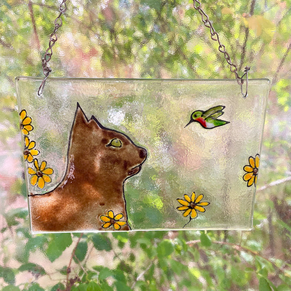 Load image into Gallery viewer, Decorative glass panel with a cat, hummingbird, and flowers design, hanging against a natural background.
