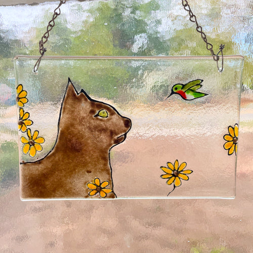 A fused glass tile hangs in a window, and features a brown cat with a green hummingbird.