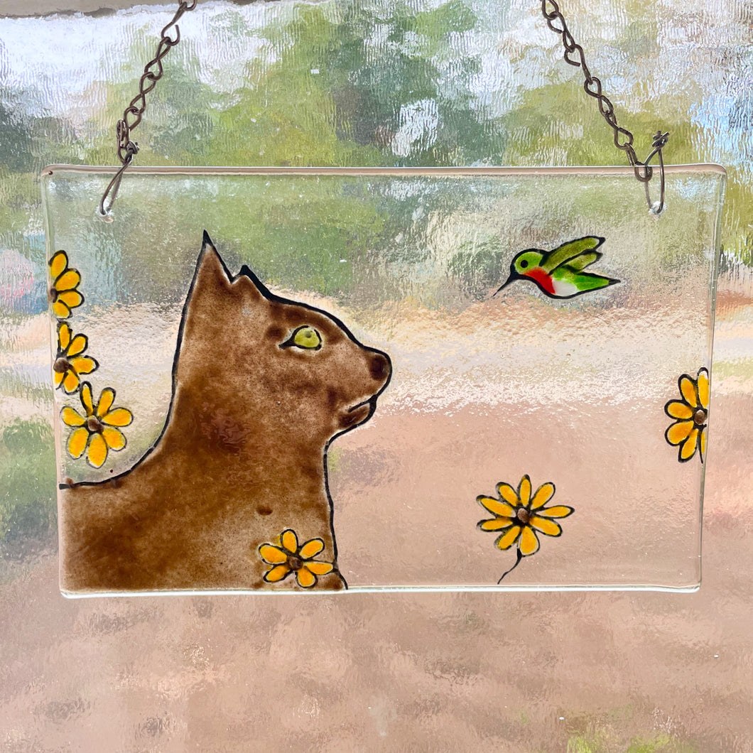 A fused glass tile hangs in a window, and features a brown cat with a green hummingbird.