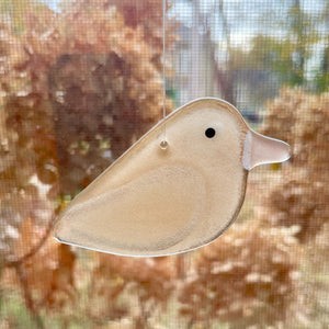 A champagne peachy pink glass bird hanging in a window. In the background are some dried hydrangea blossoms.