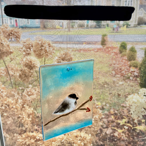 A fused glass sun catcher featuring a chickadee perching on a branch. The branch has red berries and the sky on the sun catcher is blue. The sun catcher hangs from driftwood.