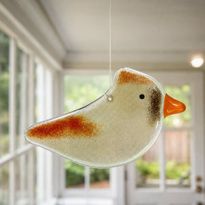 A hanging female cardinal bird-shaped glass ornament with a speckled cream body, rust-orange wing and head markings, and an orange beak, suspended indoors in front of softly blurred windows.