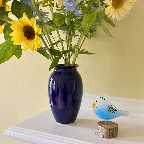 Load image into Gallery viewer, Navy blue vase with sunflowers and other flowers on a white surface with a yellow wall background. Beside the vase of flowers is a fused glass budgie made of blue and white glass. It&#39;s perched on a log slice.
