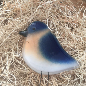 A glass bluebird with a deep blue bird with a pale flesh chest and white stomach lays on top of  nesting material