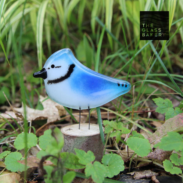 Charger l&#39;image dans la galerie, A small blue and white glass blue jay bird sits on a log in amongst green foliage.
