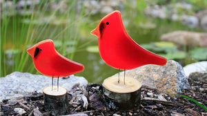 two red fused glass cardinals of different sizes are sitting on logs in front of a pond