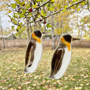 Two glass penguin ornaments hanging from a tree with a garden background.