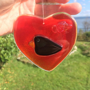 A Red Fused Glass Heart with Blackbird hangs in front of a nature scene