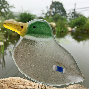 Fused glass brown and green duck ornament on a log with a pond in the background.