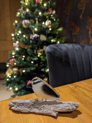 A cute fused glass nuthatch sits on a piece of driftwood on a table. In the background is a Christmas Tree