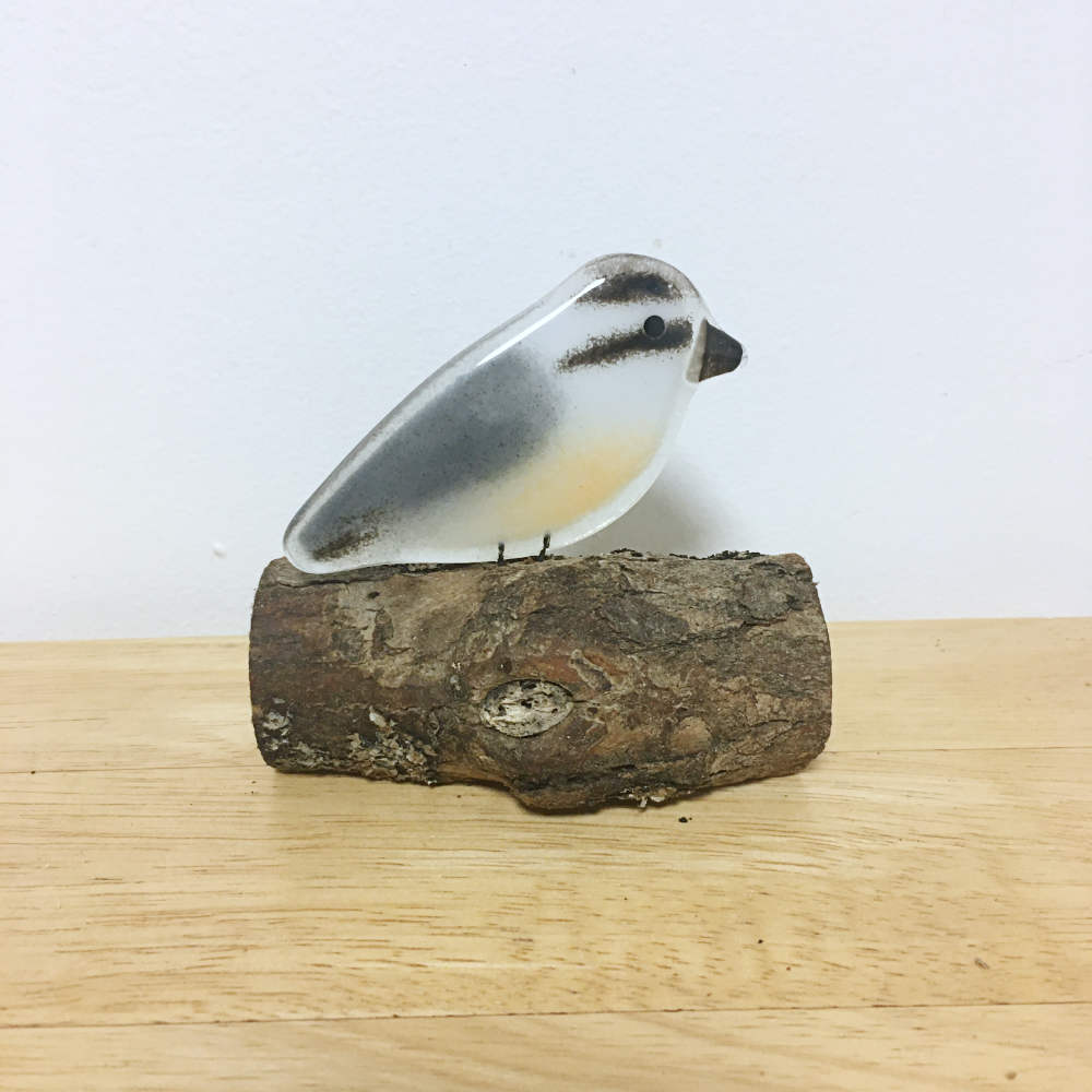 A fused glass nuthatch bird perches on a log. The bird is on a table.