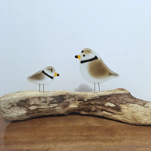 two brown and white glass birds with a black neck band perch on top of a driftwood stick