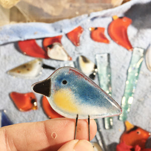 A tiny blue terracotta and white glass bird is being held between the artists fingers by its wire legs. In the background are other glass birds such as red cardinals