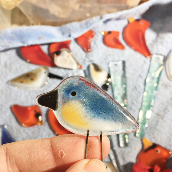 Charger l&#39;image dans la galerie, A tiny blue terracotta and white glass bird is being held between the artists fingers by its wire legs. In the background are other glass birds such as red cardinals
