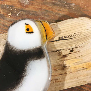 A close up of a glass puffin on a table. The puffin is laying on its side on top of some driftwood that has been branded with The Glass Bakery logo