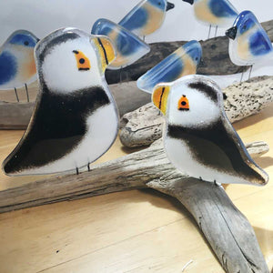 A pair of glass Puffins, one small and one large sit on a driftwood stick on a table. Behind them are glass bluebirds.