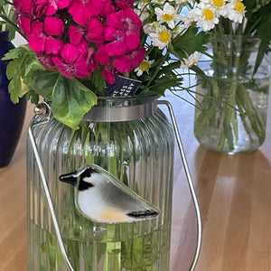 A fused glass grey and black hanging chickadee chick is suspended from flower stems in a vase of flowers.