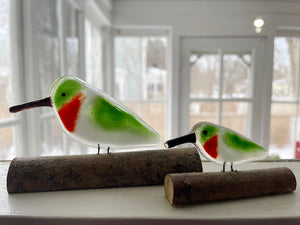 Two glass hummingbird figurines on wooden logs with a blurred indoor background