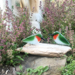 A pair of glass ruby throated hummingbirds are perched on a piece of driftwood. In the background are some dogs legs. The dog has white socks and long fur.