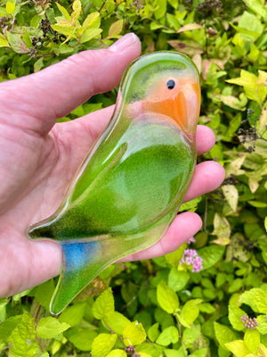 A hand holds a glossy, bird-shaped glass ornament resembling a lovebird, featuring green, orange, blue, and clear glass colors, set against a background of green foliage. The bird is being held in the hand for scale.