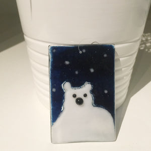 A close up of a glass polar bear tile. The tile is midnight blue with a white bear. The tile is leaning against a white planter.