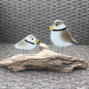 Perched pair of piping plovers made from fused glass, standing on driftwood. The background is wicker.