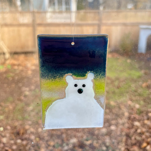 Load image into Gallery viewer, A fused glass hanging ornament featuring a white polar bear with beady eyes. The background on the tile is a midnight blue sky with a lemon coloured aurora borealis. 
