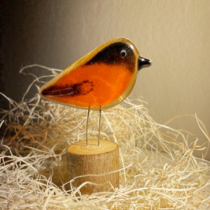 A decorative bird sculpture with a glossy orange body, black head and wing details, and thin metal legs, displayed on a small round wooden base among shredded paper against a warm beige background.