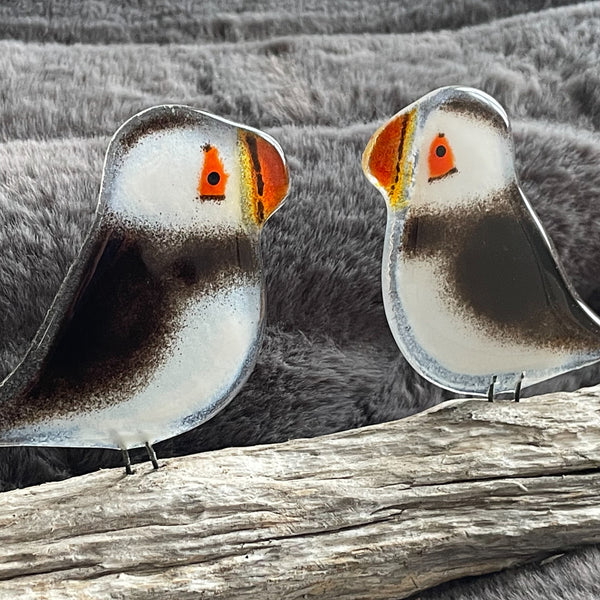 Load image into Gallery viewer, Two glass puffin bird figurines with orange beaks on a driftwood perch, in front of a warm, cozy blanket.
