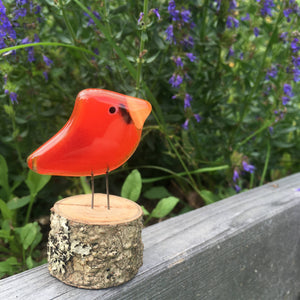 A small red glass bird in the form of a cardinal is perched on a log on the top rail of a fence. In the background are purple hyssop flowers.