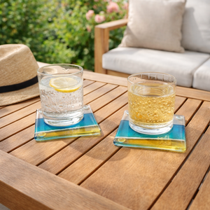 Two glasses of lemon water and sparkling water on blue and gold fused glass coasters on a wooden table outdoors.