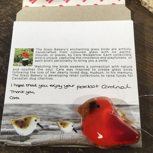 a tiny red fused glass cardinal bird sits in front of a close up of The Glass Bakery branded bird envelope