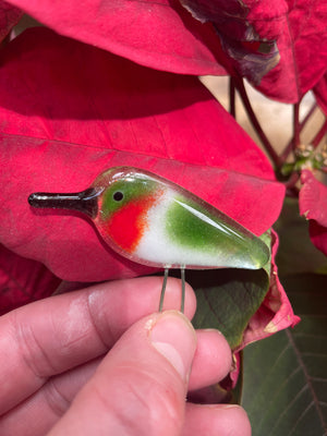 A tiny glass art ruby throated hummingbird is being held in the finger tips near to a Poinsettia Plant