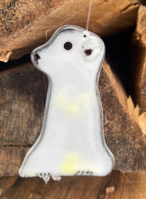 A white fused glass polar bear cub ornament hanging from clear thread in front of some logs.