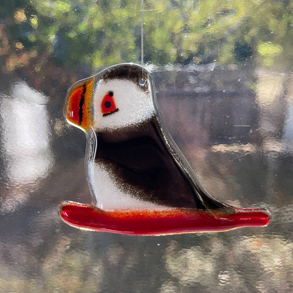 Load image into Gallery viewer, Fused Glass ornament of a puffin on a sled against blurred natural background
