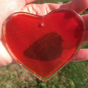 A red fused glass heart with a blackbird in the centre is being held up in a hand with the backside to the camera