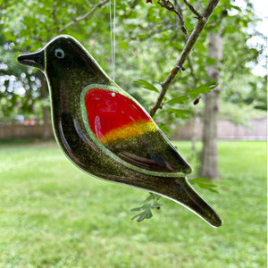 Decorative red winged blackbird glass bird with a red wing hanging from a tree branch outdoors.