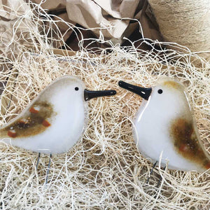 Two brown speckled and white glass birds with long black beaks, shaped to look like sandpipers. The two birds have been placed on wood fibre nesting material.