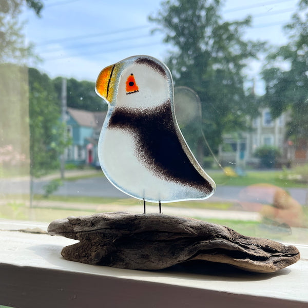 Load image into Gallery viewer, A black and white fused glass puffin is perched on driftwood, on a sash window sill.
