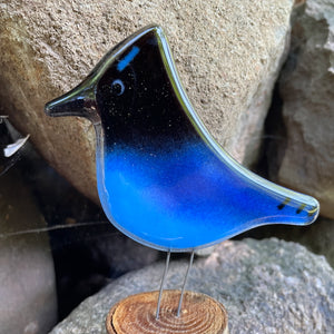 Glass bird sculpture with blue and black gradient on a wooden stand against a stone background